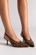 Brown high-heeled shoes with decorative elements worn by a person on a light gray background