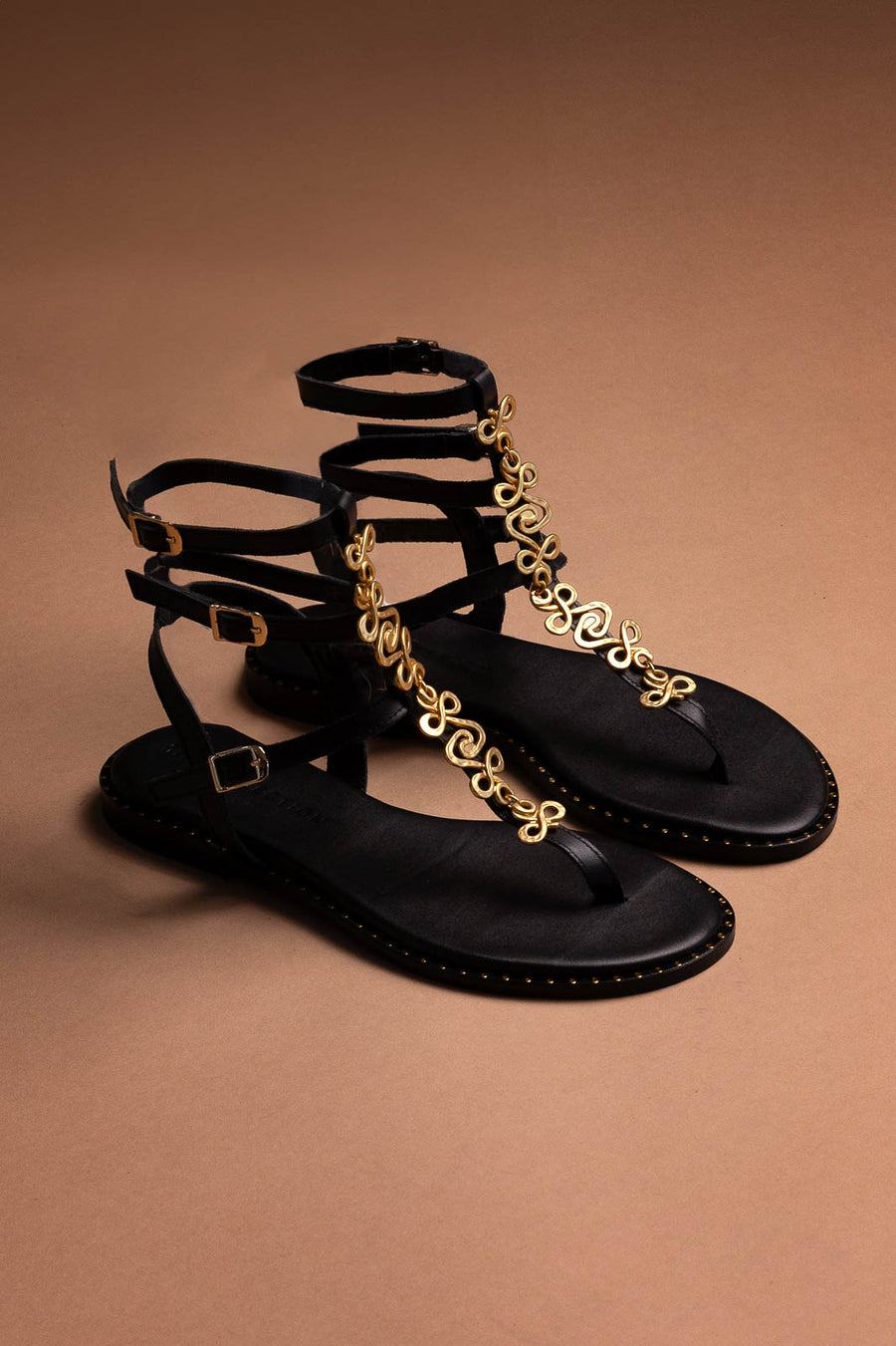 Black sandals with gold chain details on a brown background